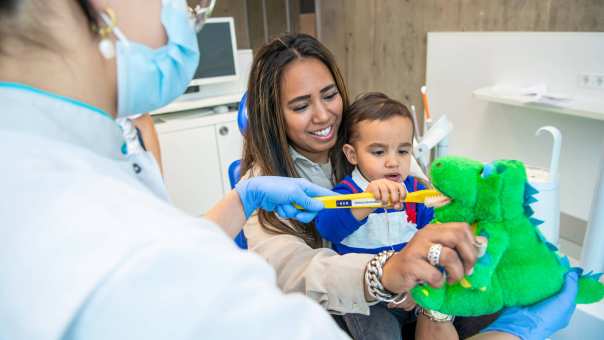 Moeder en kind zijn in de behandelruimte, met behulp van een dinosaurusknuffel leert het kind tandenpoetsen.