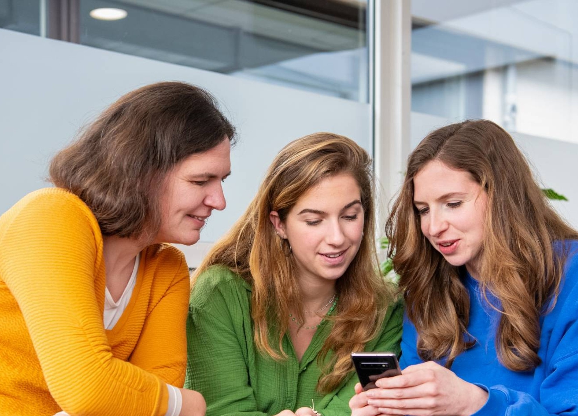 3 Studenten zitten samen op krukjes en kijken op een telefoon.