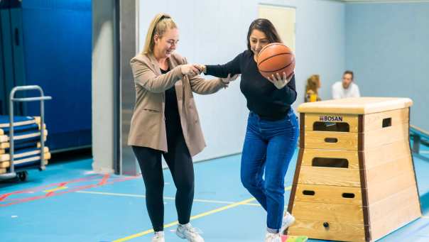 Twee studenten waarbij één student op een skateboard staat met een bal in de hand en de andere student haar begeleidt