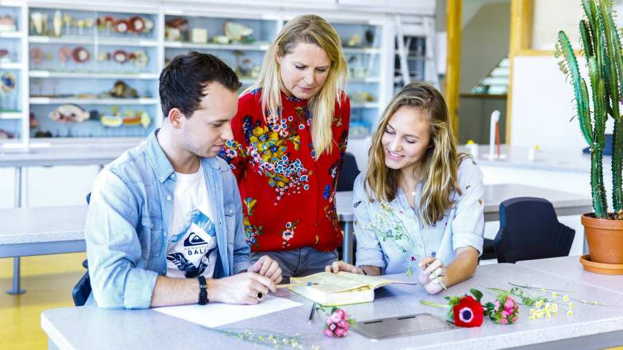 Twee studenten maken een opdracht en vragen daar iets over aan de docent die kritisch nadenkt. 