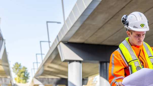 Man met veiligheidshelm en oranje werkjas checkt  de bouwtekening onder een viaduct in aanbouw.