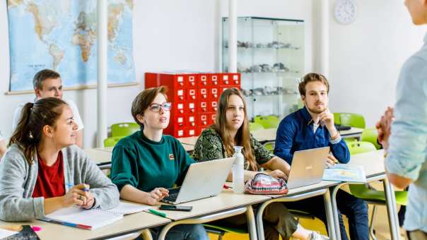 5 studenten zitten aan tafel en kijken en luisteren naar de uitleg van de docent voor hun