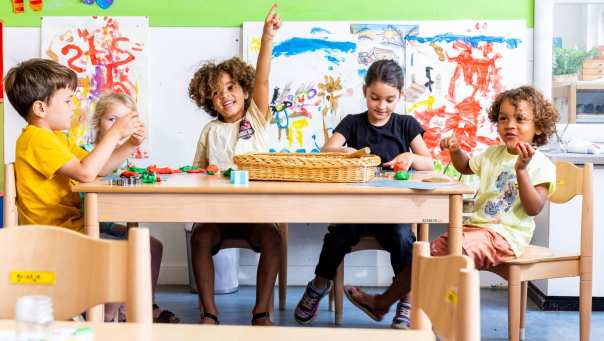 Vier kinderen zitten aan hun tafel in de klas. Een van hen steekt zijn vinger op om een vraag te stellen