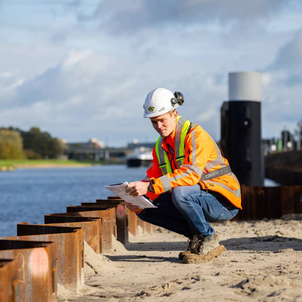 man met veiligheidsklaeding zit gehurkt bij een damwand aan een kanaal