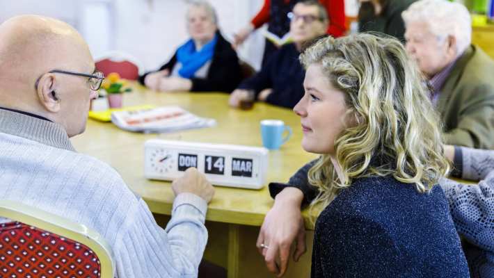 bijeenkomst wijkatelier lindenholt- aan tafel studente hurkt en geeft uitleg kalenderklok