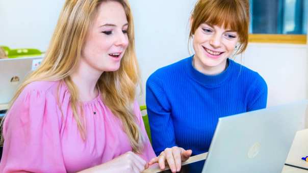 Twee vrouwelijke studenten leraar Geschiedenis zitten naast elkaar en kijken lachend naar het scherm (laptop) dat voor hun staat.