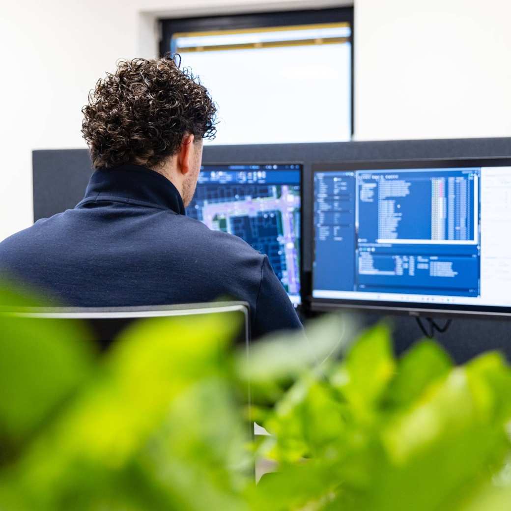 Man werkt op twee beeldschermen aan een bouwontwerp. Op de voorgrond staat een plant