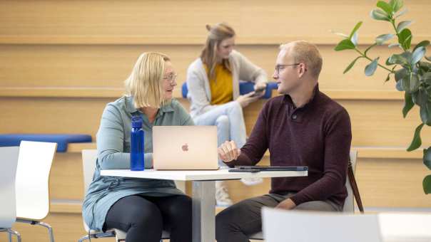 2 studenten aan het overleggen, 1 student zit op de achtergrond op de trap op haar mobiel.