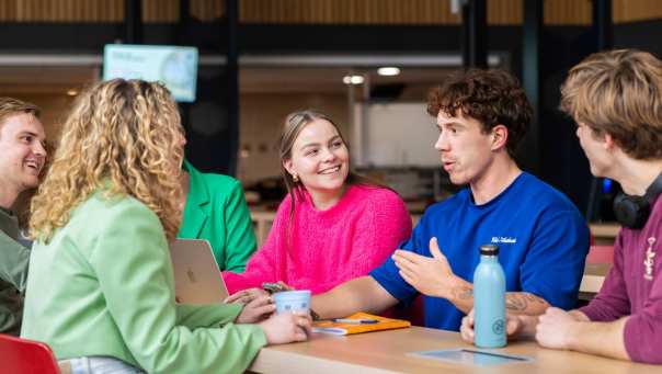 Groep studenten zit aan een lange tafel, lachen en kletsen samen.