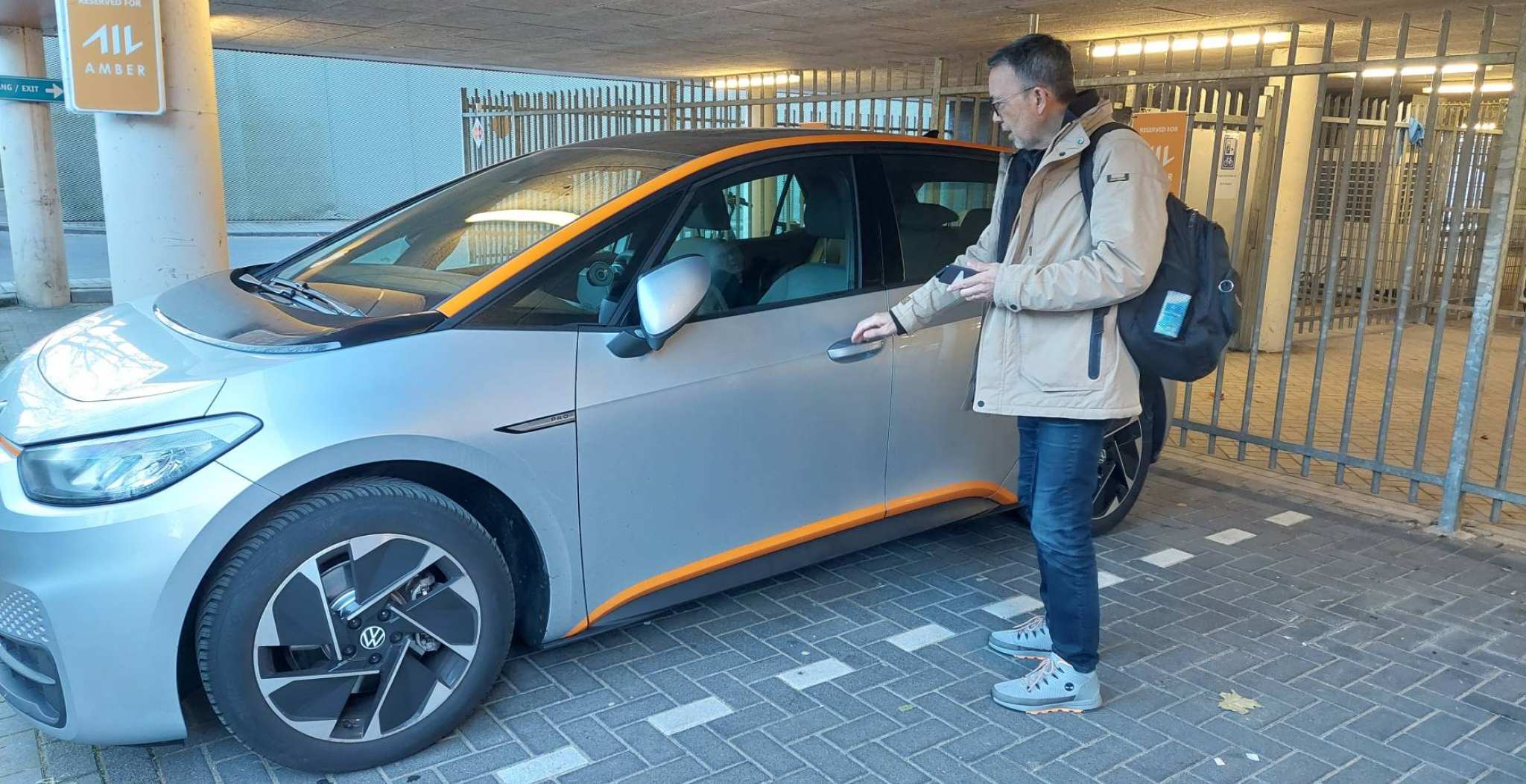 Docent zorg en techniek Clark Nowack opent de grijze elektrische Volkswagen van Amber met 1 hand op de klink en in de andere een smartphone voor de app.