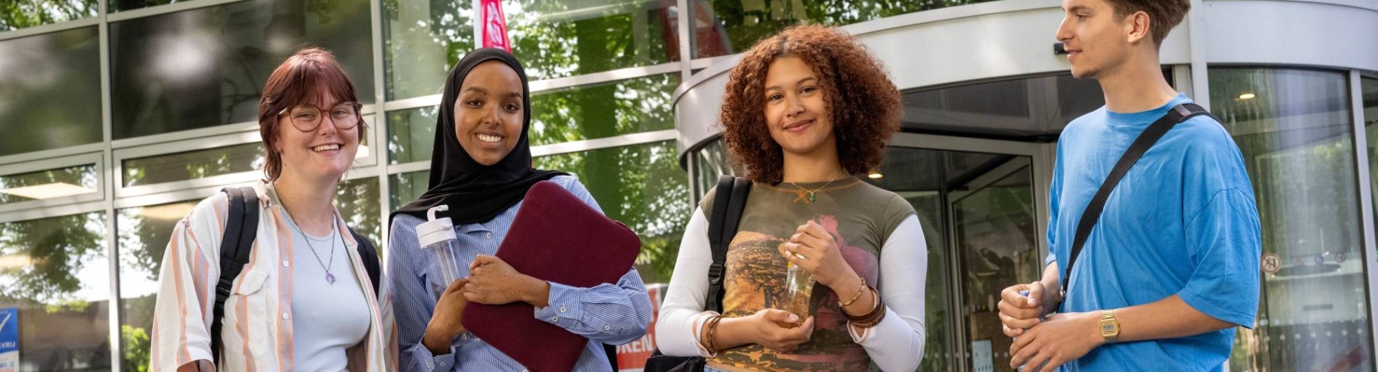 Vier studenten staan bij gebouw LvS10 bij draaideur, buiten. Op een rijtje. 