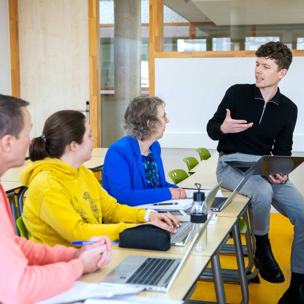 een masterstudent zit op de rand van de tafel met een laptop in z'n hand en legt wat uit aan de rest