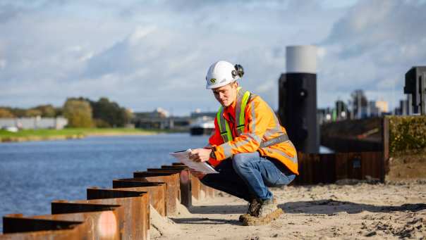 Man met veiligheidshelm en oranje werkjas zit gehurkt bij een damwand aan een kanaal.