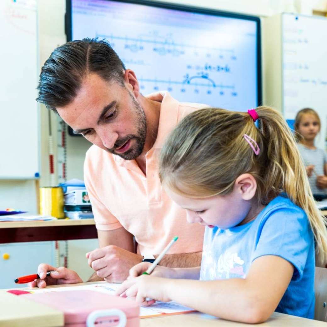 Een student aan de pabo helpt een meisje aan tafel in de klas.