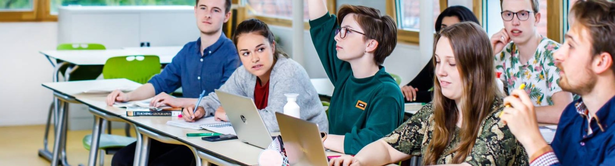 Leraar staat les te geven voor de klas en geeft de beurt aan een leerling die vooraan zit met zijn vinger omhoog.