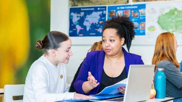 Docente Angel Olatunji legt wat uit aan een leerlinge en gebruikt daarbij gebaren, een schrift en een laptop, op het Citadel College. 