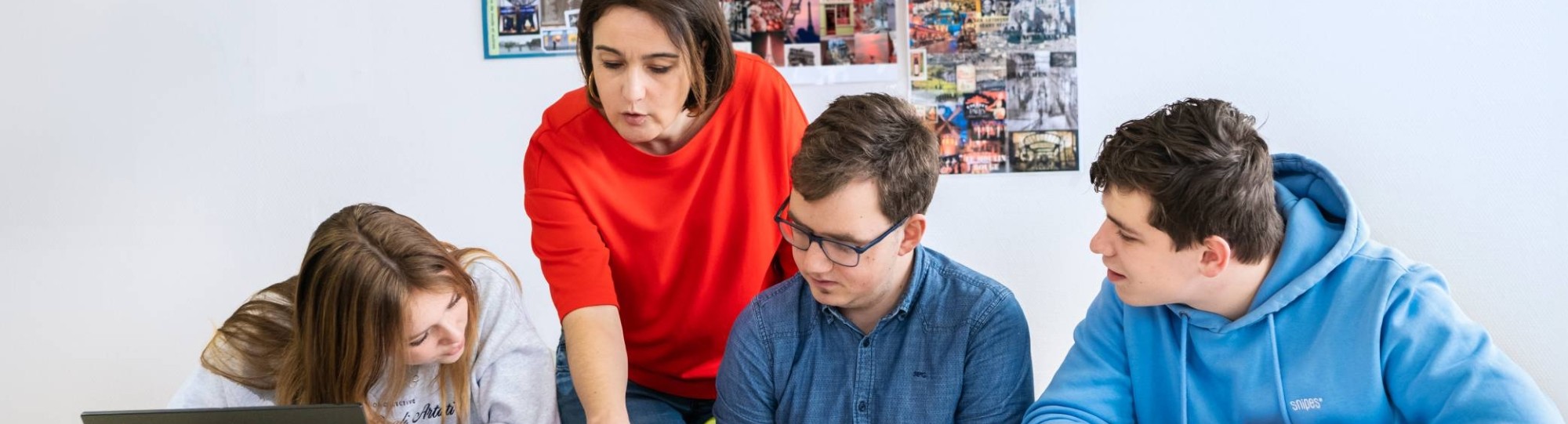 Drie studenten zitten op een rij en de docent staat achter hen. Ze wijst in het boek en de studenten kijken mee.