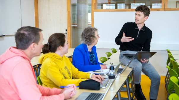 masterstudent legt wat uit aan de anderen terwijl hij op een bureau zit met een laptop op schoot