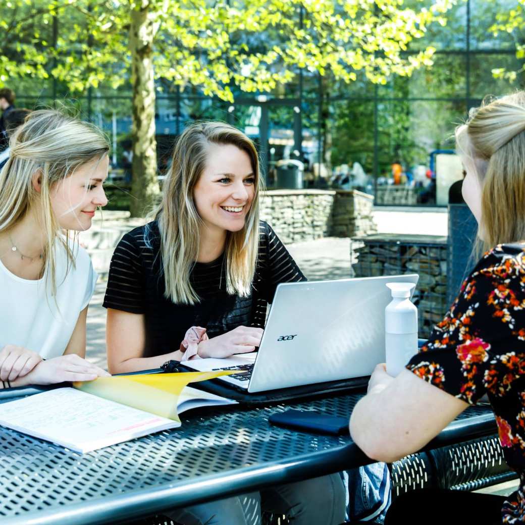 Drie studenten zitten buiten op het HAN-terrein te werken met hun laptops.