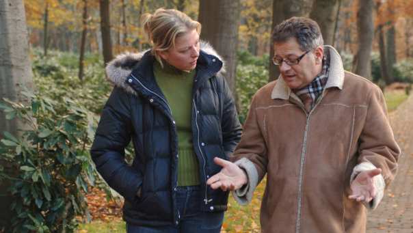 Een oudere heer en een zorgmedewerker overleggen, wandelend in het bos.