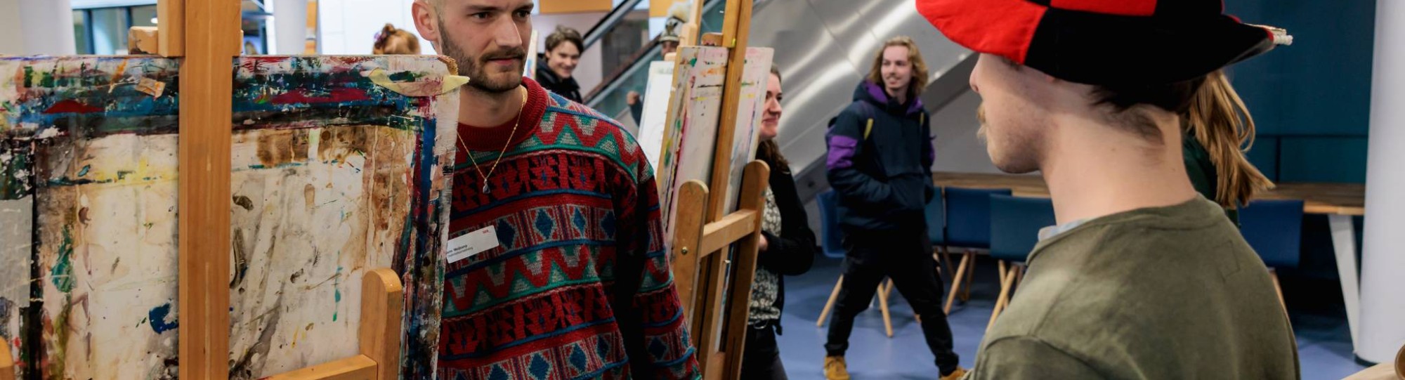 Twee mannen waarvan een met een zwart rode hoed staan elkaar bij schildersezels te observeren