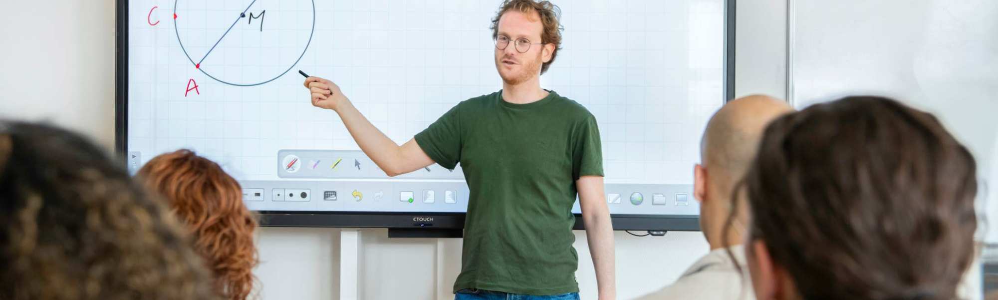 Leraar Wiskunde kijkt naar zijn klas, terwijl zijn hand wijst naar de cirkel op het bord waar 'stelling van Thales' boven staat
