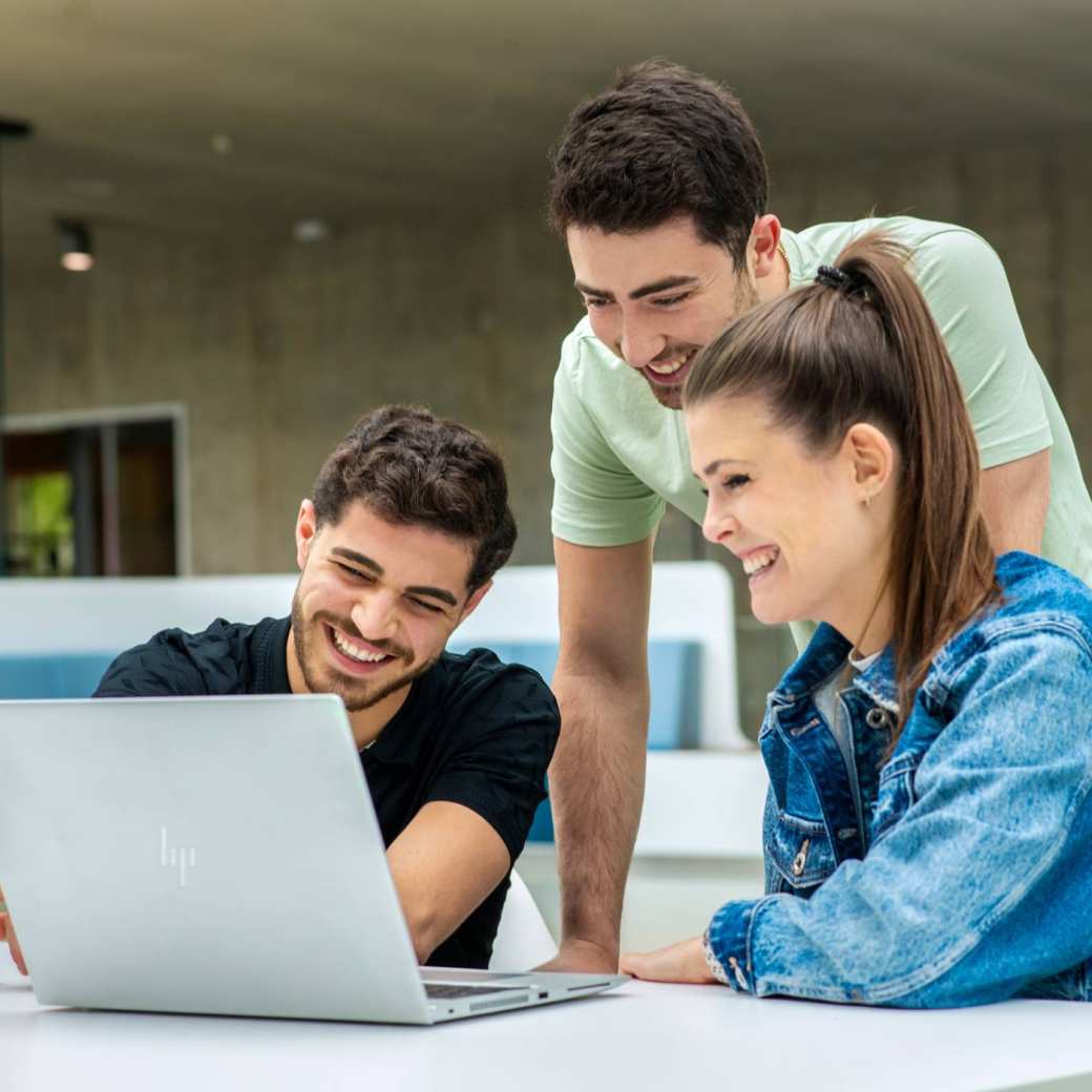 2 studenten zitten aan tafel met 1 laptop, een klasgenoot staat erachter en lacht.