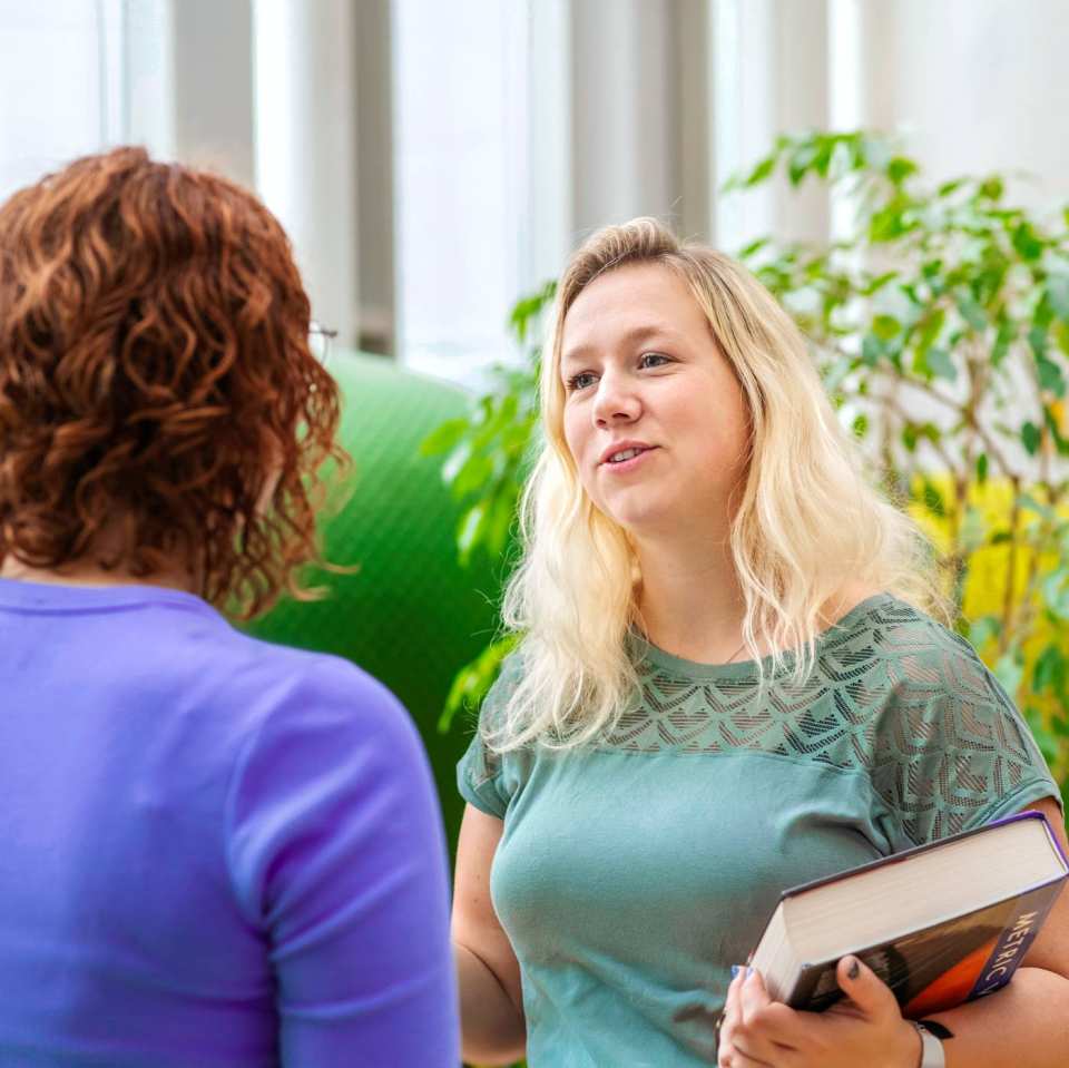 Student Leraar Wiskunde heeft boek vast en praat tegen een medestudent. Ze staan tegenover elkaar
