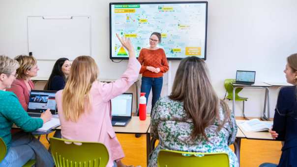 Docent staat voor een volle klas en kijkt lachend naar de student die de vinger opsteekt. 