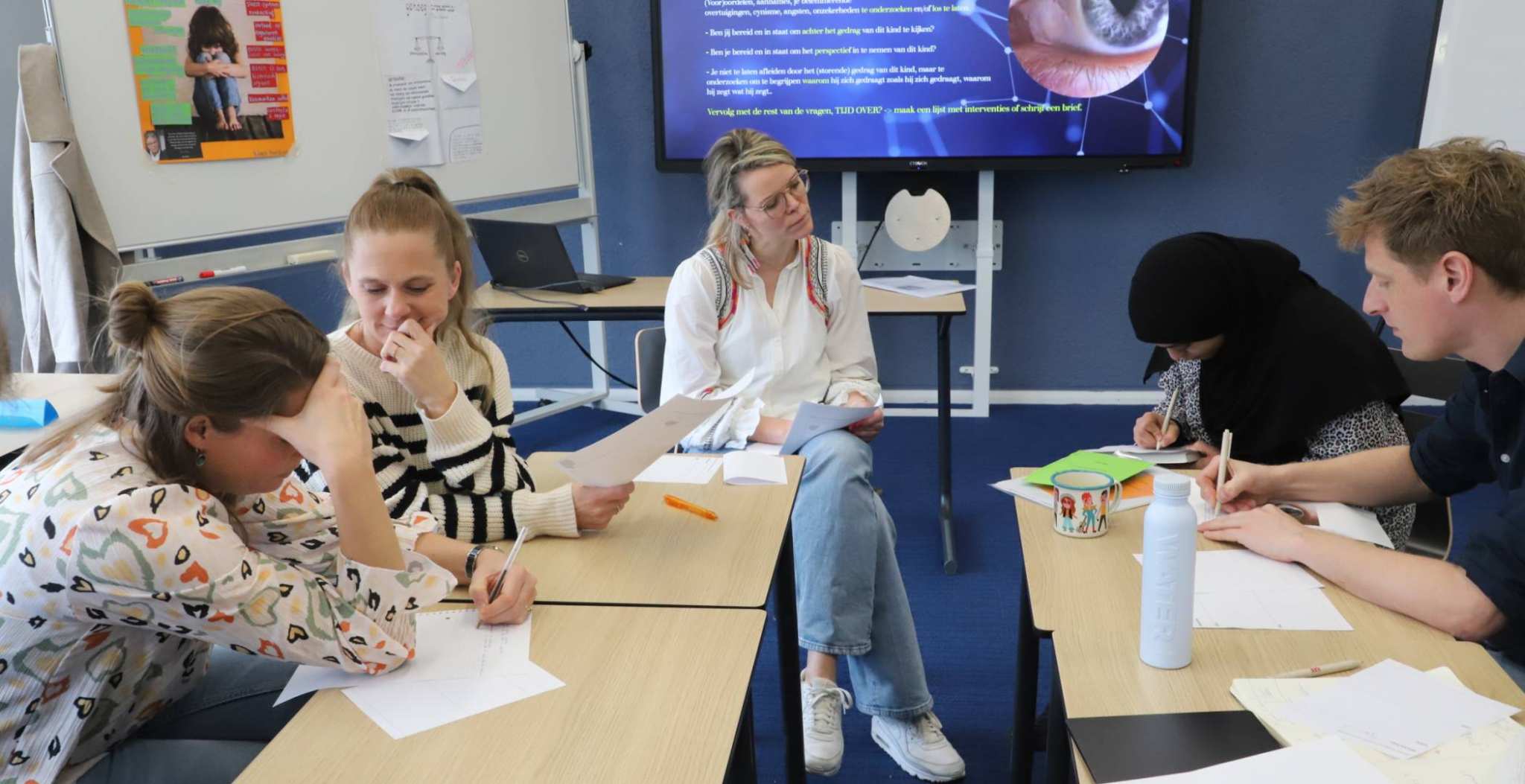 Jenna Gudden zit voor de groep. Ze heeft blond haar, draagt een bril en een witte blouse met een lichtblauwe spijekrbroek. Voor haar zitten medestudenten en docenten te werken aan een opdracht.