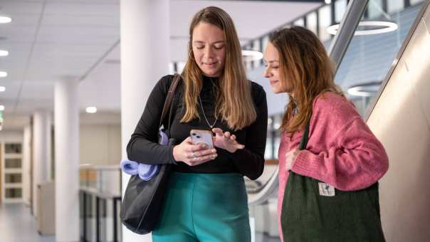 2 studenten met tas kijken op mobiel in de gang.