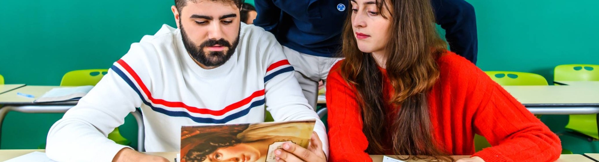 Twee studenten kijken naar een historisch boek en de docent kijkt observerend mee. 