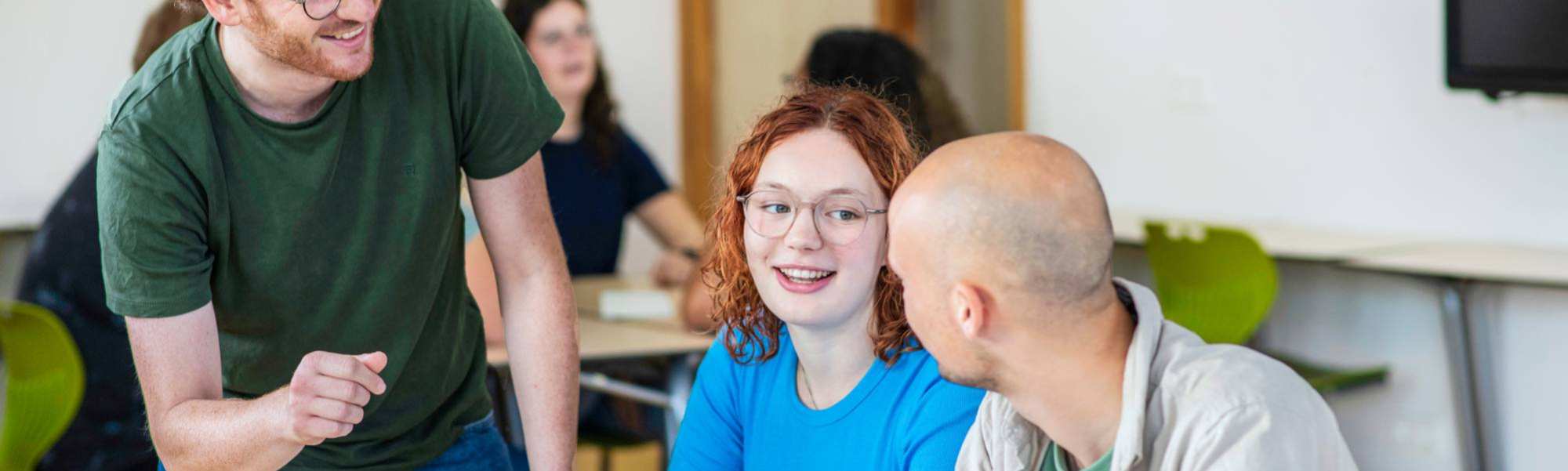 Leraar Wiskunde luistert lachend naar zijn twee studenten die iets zeggen tegen hem
