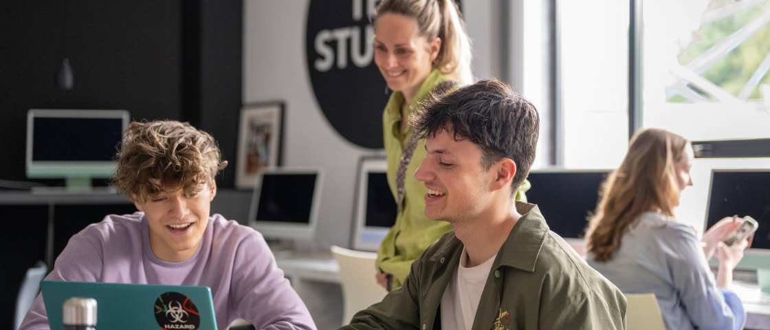 Twee studenten in een klas overleggen samen, docent kijkt mee. 