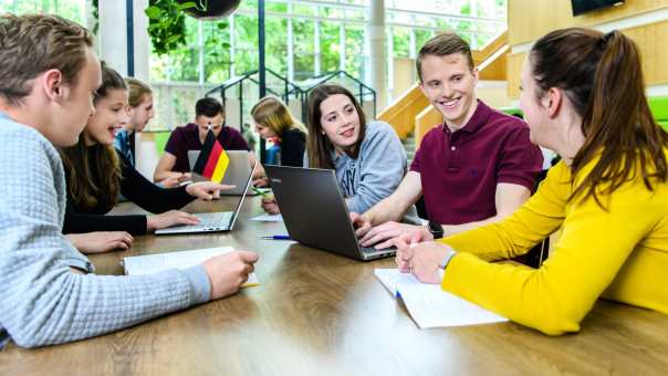 Studenten leraar Duits zitten aan een grote tafel in de kantine van het I/O gebouw en zijn aan het overleggen. 