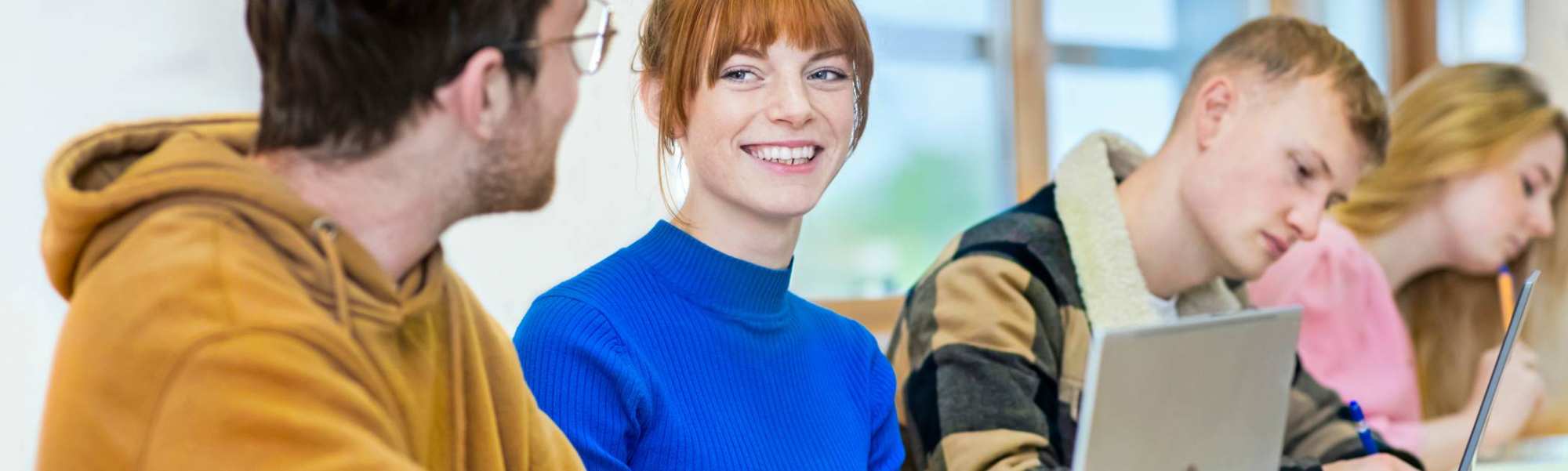 Vier studenten zitten op een rij. Een student specifiek is scherp in beeld en ze lacht naar de persoon naast haar.