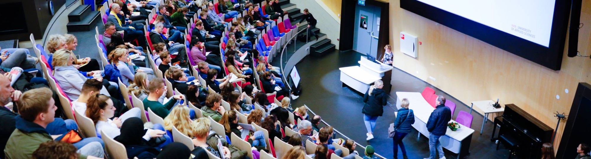 Beeld van bovenaf in auditorium met volle zaal voor presentatie