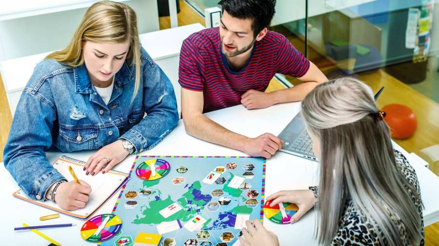 Drie studenten Leraar Frans spelen aan de tafel een bordspel bestaande uit allerlei kleuren. Eén meisje houdt de scores bij. 