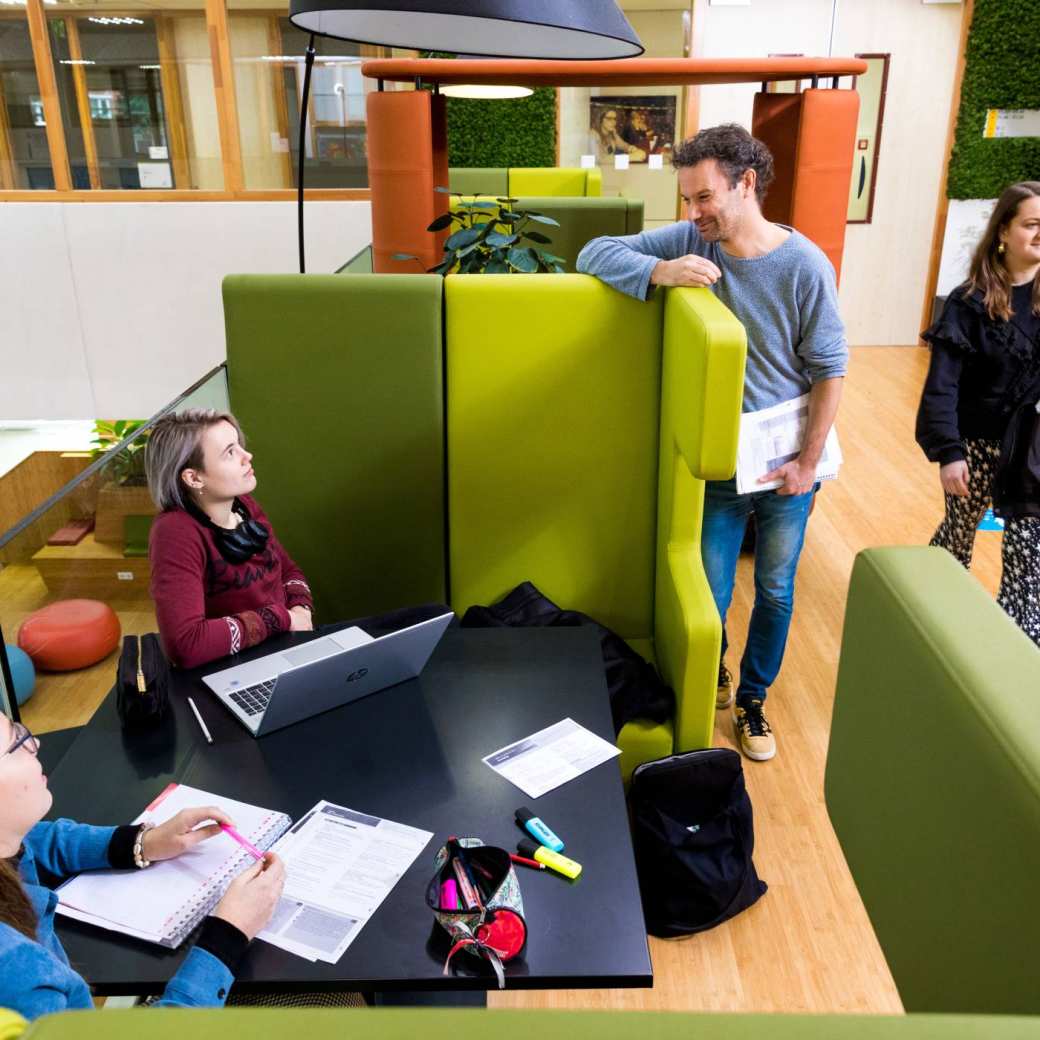 Leraar spreekt twee studenten aan die zitten te werken aan de tafel in het I/O gebouw.