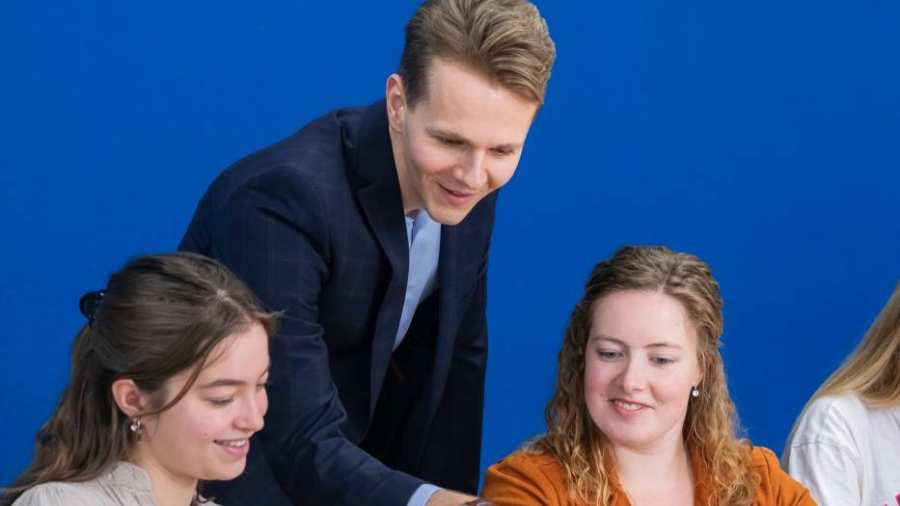 Docent in donkerblauw pak kijkt mee op de laptop bij twee van zijn studenten.
