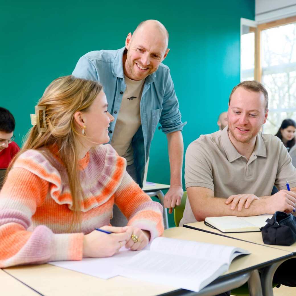 Drie studenten zitten rond de tafel in een lokaal, terwijl de docent uitleg geeft en naar het boek op de tafel wijst.