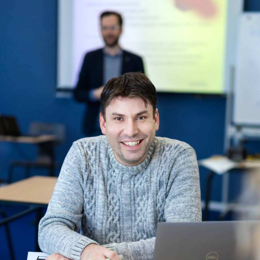 ALLEEN VOOR DE MANP | Een masterstudent met laptop bij een les onderzoek met op de achtergrond een docent van de Master Advanced Nursing Practice.