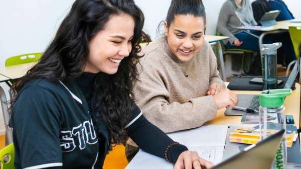 Twee studenten werken samen op één laptop en lachen naar het scherm.
