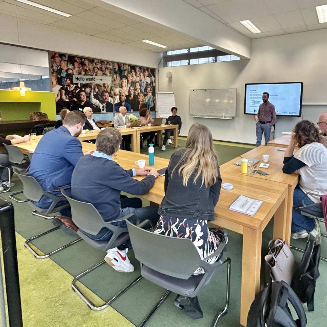 Ongeveer 20 mensen zitten in een U vorm aan tafels, te luisteren naar een presentatie