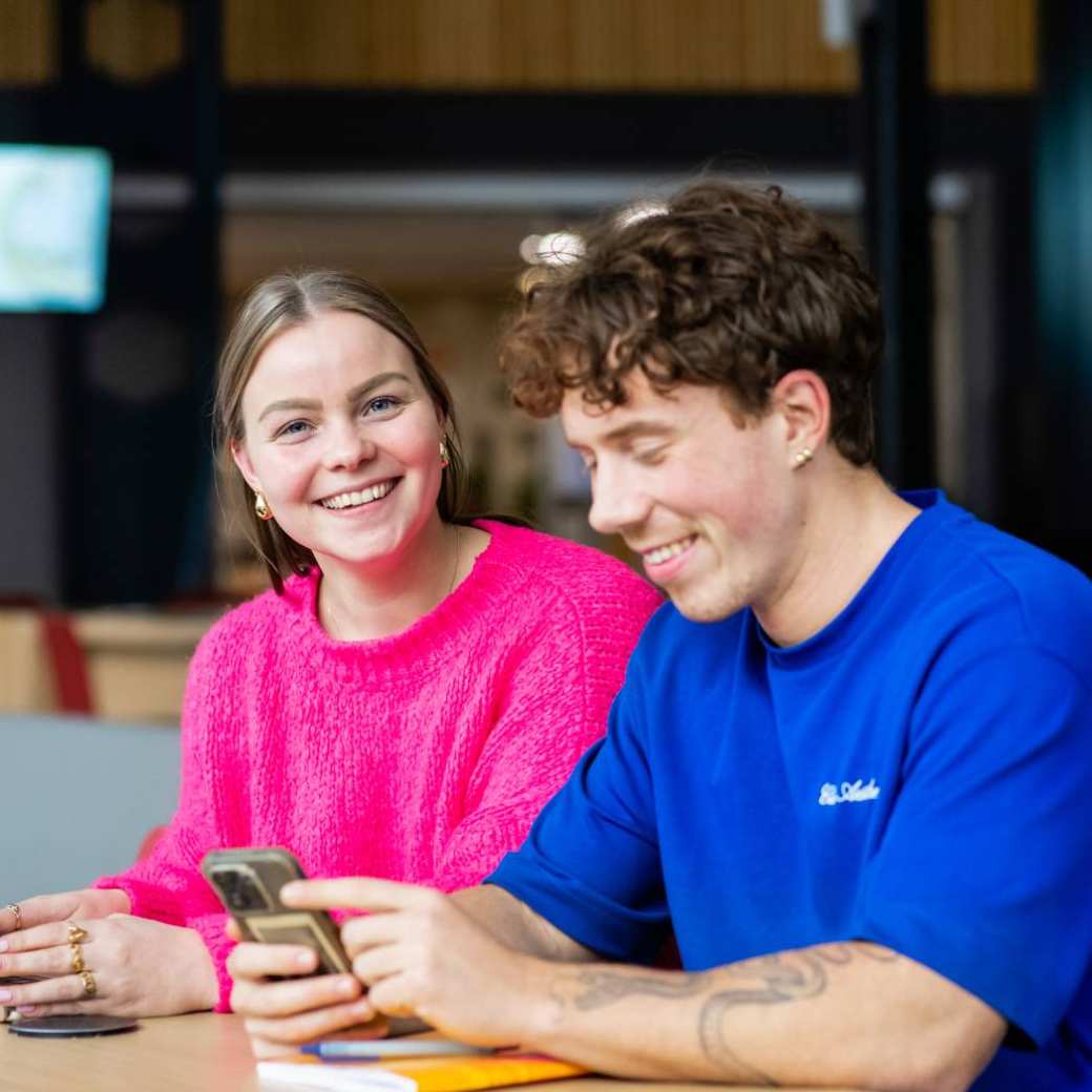 2 studenten aan een tafel. 1 kijkt lachend in de camera, de ander kijkt naar zijn telefoon.