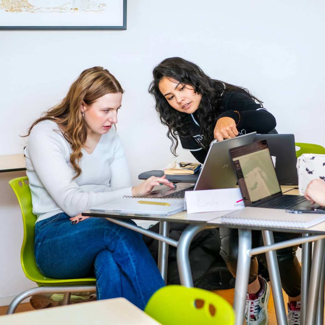 Docent geeft aan twee studenten uitleg en wijst daarbij naar de pagina in het boek, dat de studenten in de hand hebben.