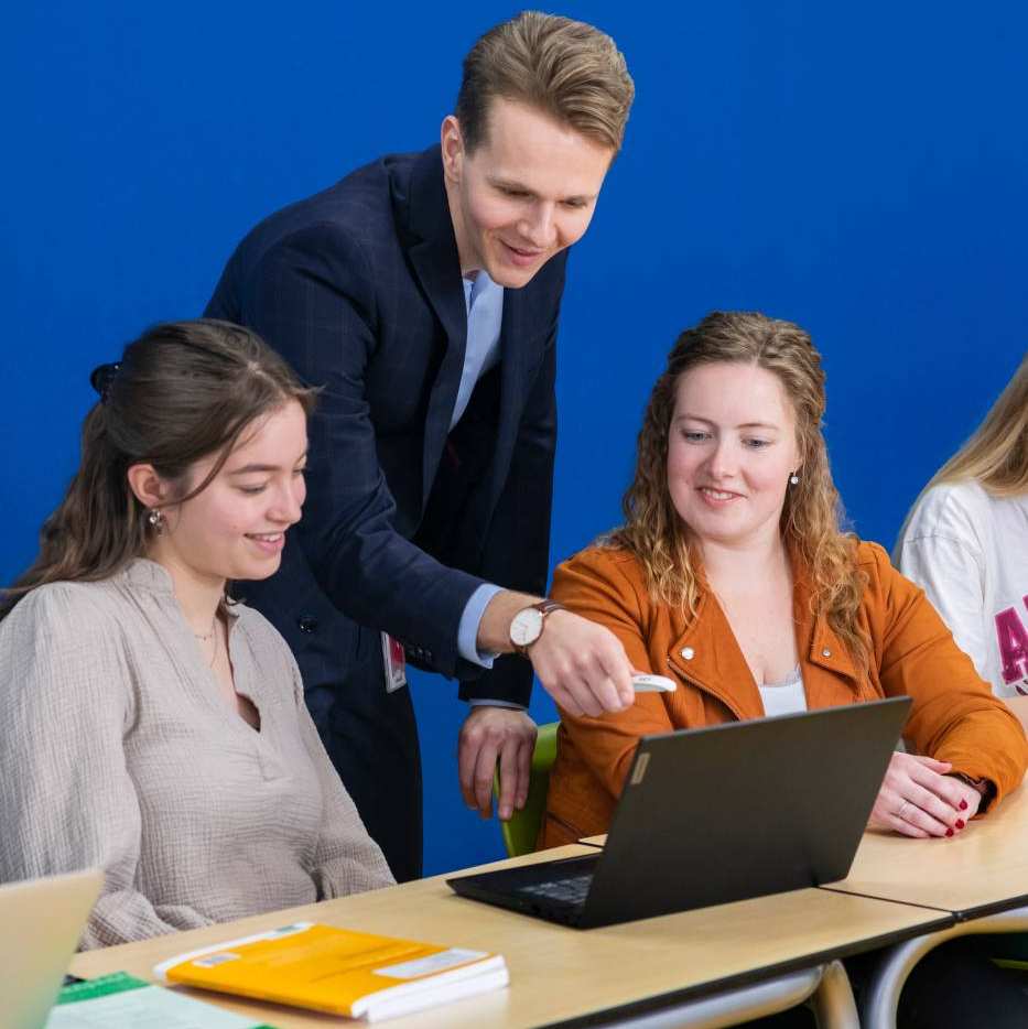 Docent in donkerblauw pak kijkt mee op de laptop bij twee van zijn studenten.
