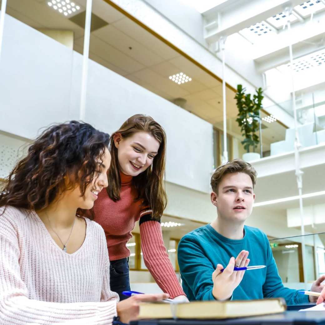 Drie studenten zitten te werken en één student stelt een vraag aan de docent die aandachtig luistert.