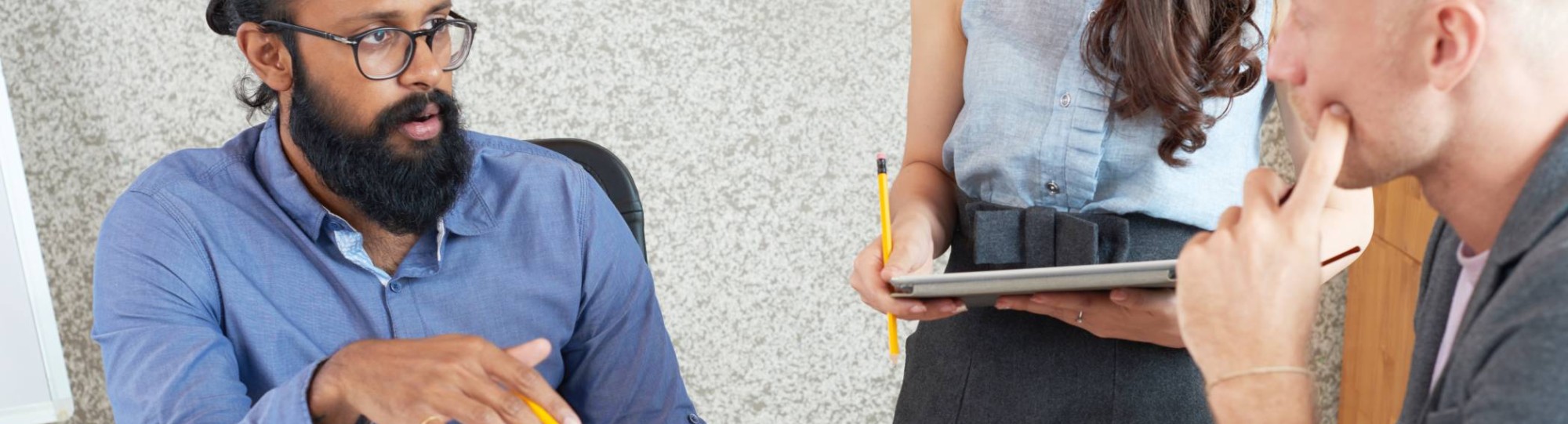 Man in een blauwe blouse legt iets uit aan een vrouw en een man die samen aan een tafel zitten met een laptop. Samenwerken. Overleg. Seece.