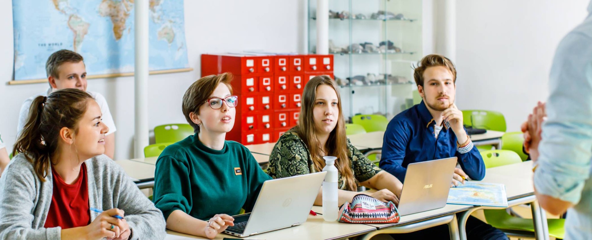 5 studenten zitten aan tafel en kijken en luisteren naar de uitleg van de docent voor hun